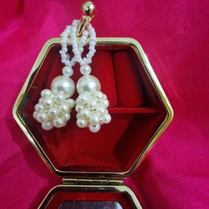 May include: A pair of white beaded tassel earrings displayed in an open, red velvet-lined, gold-trimmed jewelry box. The earrings feature large, round pearl-like beads and smaller seed beads. The background is a vibrant pink fabric.