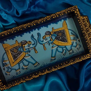 May include: Rectangular wooden tray with a hand-painted design of two blue elephants with gold and yellow accents. The tray has a dark border with gold decorative trim and handles. The background is a blue satin fabric.