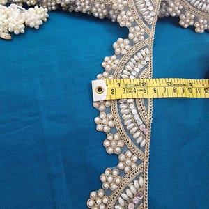 May include: Close-up of decorative trim with pearls and mirrored accents. The trim is measured with a yellow measuring tape, showing a width of approximately 2 inches. The trim is on a teal blue fabric.