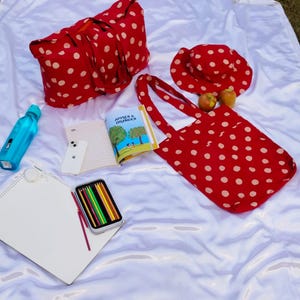 May include: A red tote bag, hat, and travel bag with white polka dots are arranged on a white surface. A book titled "Apples & Oranges" is open next to a smartphone, colored pencils, and a water bottle. Two apples are also present.