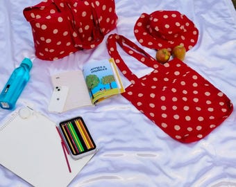 May include: A red tote bag, hat, and travel bag with white polka dots are arranged on a white surface. A book titled "Apples & Oranges" is open next to a smartphone, colored pencils, and a water bottle. Two apples are also present.