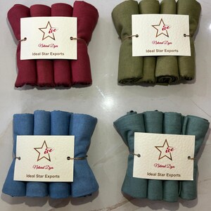 May include: Four bundles of rolled fabric in shades of burgundy, olive green, blue, and teal. Each bundle is tied with twine and has a white tag with a star logo and the text "Natural Dyes" and "Ideal Star Exports".