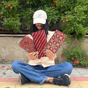 May include: Three red and patterned fabric pouches are held up in front of a person. The pouches have different designs, including stripes and floral patterns. The person is wearing a white cap, a beige shirt, and blue jeans.