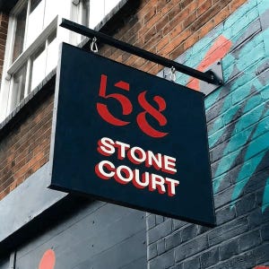 May include: A rectangular sign with the number "58" in red and the words "STONE COURT" in white, hanging from a black metal bracket. The sign is against a brick wall.