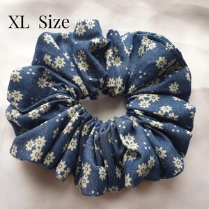 May include: XL size denim scrunchie with a white floral pattern. The hair accessory is made of gathered fabric, creating a voluminous, circular shape. The scrunchie is a deep blue colour with small white flowers.