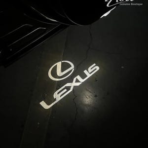 May include: A white Lexus logo and wordmark projected onto a dark surface from a vehicle door light. The projection displays the circular Lexus emblem above the brand name. The text Abel Genuine Boutique is visible in the upper right corner.