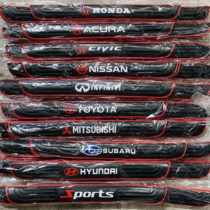 Op de afbeelding: Een verzameling zwarte dorpelbeschermers voor autodeuren met rode randen, elk met een ander autologo in het wit, waaronder Honda, Acura, Civic, Nissan, Infiniti, Toyota, Mitsubishi, Subaru, Hyundai en Sports.