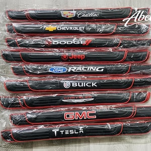 May include: Black car door sill protectors with red trim, featuring logos for Cadillac, Chevrolet, Dodge, Jeep, Ford Racing, Buick, Chrysler, GMC, and Tesla. Each protector is individually packaged.