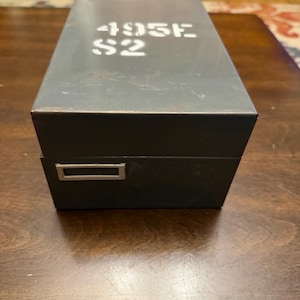 May include: A rectangular, dark gray metal storage box with a hinged lid. The top of the lid features the white stenciled text 495E S2. The front of the box includes a small, rectangular silver-colored metal label holder or pull handle.