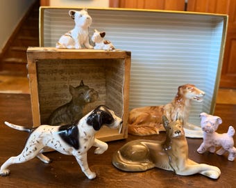 May include: A collection of seven ceramic dog figurines in various poses and breeds, including a white and black spotted pointer, a brown reclining dog, a small pink puppy, and a dark grey dog inside a wooden box with musical note patterns.