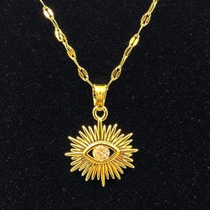 May include: Gold-colored necklace with a pendant featuring an eye design. The pendant has a sunburst pattern around a central clear stone. The chain is a delicate gold color, and the background is black.