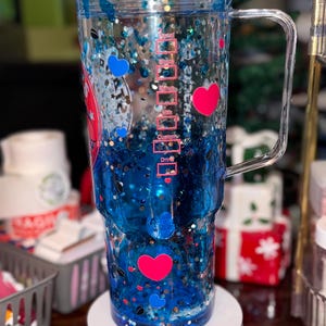 Puede incluir: Un vaso transparente de doble pared con asa, lleno de l&iacute;quido azul, purpurina y confeti en forma de coraz&oacute;n. El vaso tiene un dise&ntilde;o de coraz&oacute;n rosa y azul. El lateral del vaso tiene marcas de medici&oacute;n.