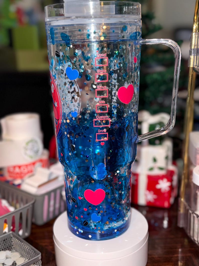 Puede incluir: Un vaso transparente de doble pared con asa, lleno de l&iacute;quido azul, purpurina y confeti en forma de coraz&oacute;n. El vaso tiene un dise&ntilde;o de coraz&oacute;n rosa y azul. El lateral del vaso tiene marcas de medici&oacute;n.