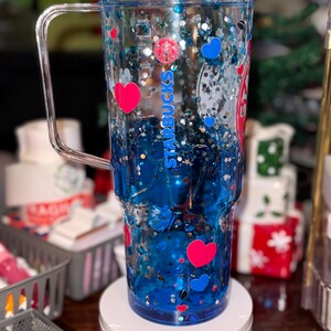 Puede incluir: Vaso transparente con asa, lleno de l&iacute;quido azul, purpurina y confeti en forma de coraz&oacute;n y redondo. El vaso tiene el logotipo y el texto de Starbucks. El dise&ntilde;o incluye corazones rojos y azules. El vaso est&aacute; sobre un soporte blanco.