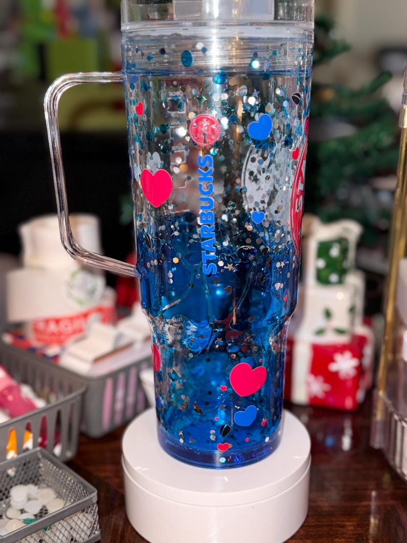 Puede incluir: Vaso transparente con asa, lleno de l&iacute;quido azul, purpurina y confeti en forma de coraz&oacute;n y redondo. El vaso tiene el logotipo y el texto de Starbucks. El dise&ntilde;o incluye corazones rojos y azules. El vaso est&aacute; sobre un soporte blanco.