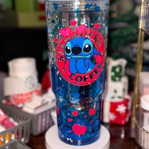 Puede incluir: Un vaso transparente con un dise&ntilde;o de purpurina azul y plateada. El vaso presenta un logotipo del personaje Stitch con corazones. La taza est&aacute; llena de l&iacute;quido azul y confeti en forma de coraz&oacute;n. El vaso est&aacute; sobre un soporte blanco.