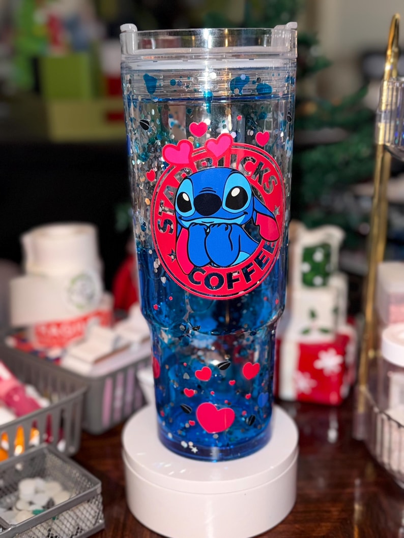 Puede incluir: Un vaso transparente con un dise&ntilde;o de purpurina azul y plateada. El vaso presenta un logotipo del personaje Stitch con corazones. La taza est&aacute; llena de l&iacute;quido azul y confeti en forma de coraz&oacute;n. El vaso est&aacute; sobre un soporte blanco.