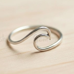 May include: A silver ring with a wave design. The ring has a curved wave shape on top, made from a shiny metal. The ring is set against a light brown wooden surface.
