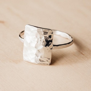 May include: A silver ring with a rectangular hammered design. The ring has a simple band and a textured, reflective surface on the rectangular face. The ring is displayed on a light wooden surface.