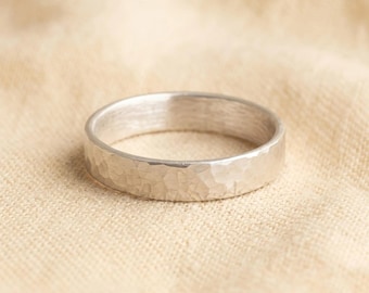 May include: A silver-colored band ring featuring a textured, hammered finish on the outer surface. The ring has a consistent width and a smooth, polished interior edge, showcasing a metallic sheen across the entire circular structure.