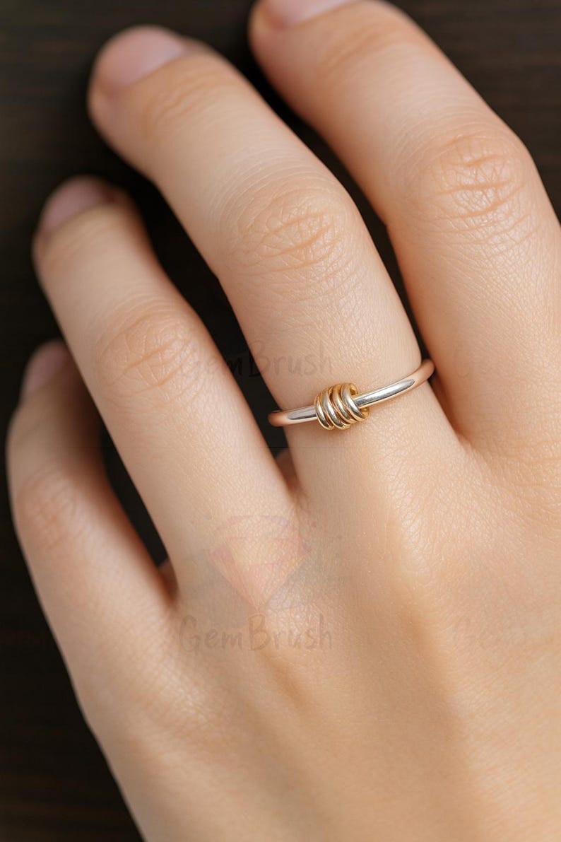 May include: A silver ring with three gold-colored interlocking circles as a focal point. The ring is on a finger, showcasing the contrast between the silver band and the gold accents. The design is simple yet elegant.
