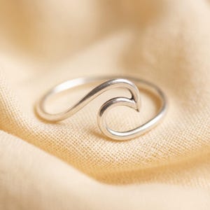 Puede incluir: Un anillo de plata pulida con una banda continua que se curva formando una onda estilizada en la parte superior. El metal tiene una superficie lisa y reflectante, y el diseño consiste en una sola pieza de alambre de plata en forma circular.