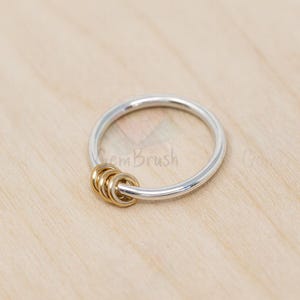 May include: A silver ring with three gold-colored interlocking rings. The ring is set against a light wood grain background. The ring is a simple, elegant design.