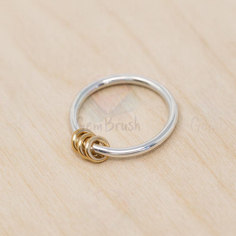 May include: A silver ring with three gold-colored interlocking rings. The ring is set against a light wood grain background. The ring is a simple, elegant design.