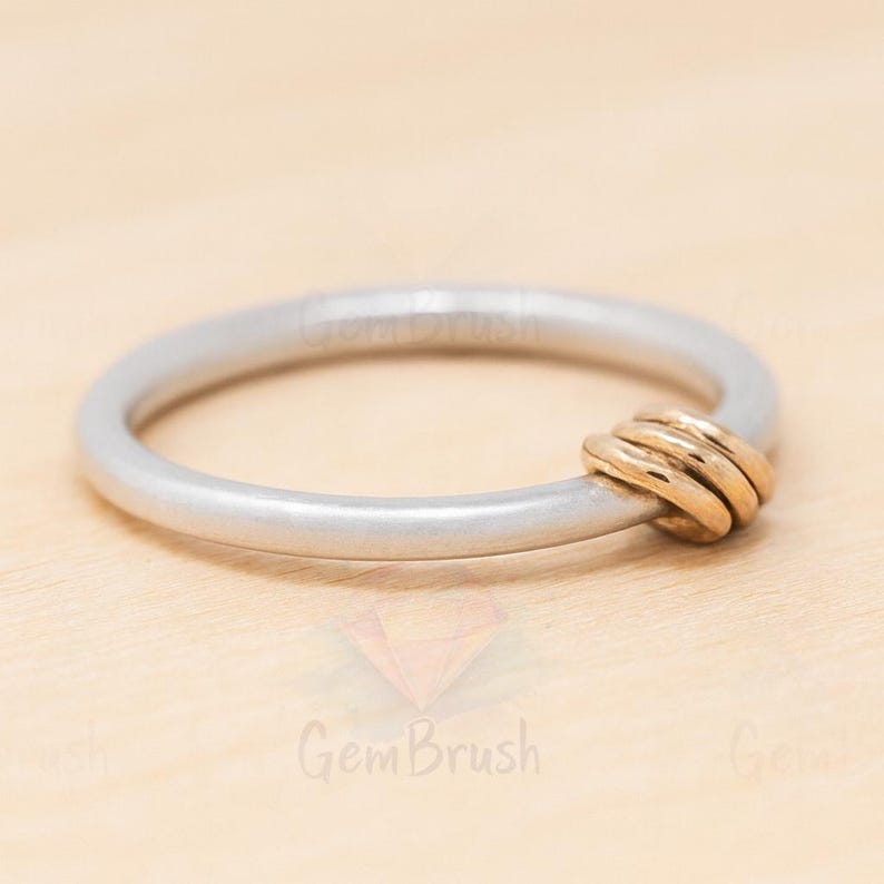 May include: A silver-colored bangle bracelet with a gold-colored knot design. The bracelet is circular and appears to be made of metal. The gold knot is on the side of the bracelet. The background is a light wood color.