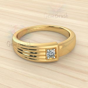 May include: A gold ring featuring a square, clear gemstone set in a square bezel. The ring band has a smooth, polished finish with three parallel grooves on one side. The ring is displayed on a light wood surface.