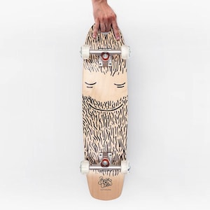 May include: A light-coloured skateboard with a whimsical beard design. The board features black lines that create the beard, moustache, and eyes. The wheels are white, and the trucks are silver and red. The brand name "Paise Ville Skateboards" is printed on the bottom.