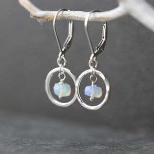 May include: A pair of silver leverback earrings. Each earring features a small, faceted, oval-shaped opal gemstone suspended within a hammered silver hoop. The earrings are displayed against a blurred gray background.