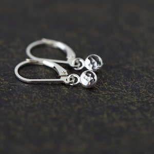 May include: A pair of silver leverback earrings. Each earring features a faceted, round charm. The earrings are displayed on a dark, textured surface. The earrings are designed to be worn by anyone.