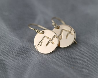 May include: A pair of gold-colored, round earrings. Each earring features a mountain range design etched into the surface. The earrings have a simple hook closure and are displayed against a gray background.