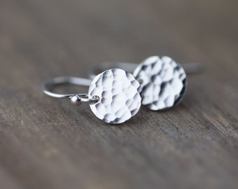 May include: A pair of silver earrings with hammered, circular pendants. The earrings have a simple hook design and a textured surface on the circular discs. The earrings are displayed on a wooden surface.