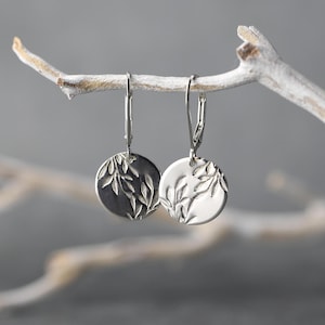 May include: A pair of silver-toned earrings with a circular design. Each earring features a detailed leaf pattern. The earrings are suspended from a silver lever-back clasp. The background is a soft, neutral gray.