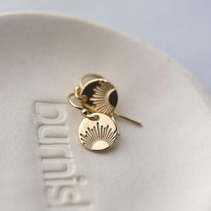 May include: A pair of gold-colored dangle earrings. Each earring features a small, round pendant with a sunburst design. The earrings are displayed on a white, textured surface, showcasing their delicate design and warm metallic hue.