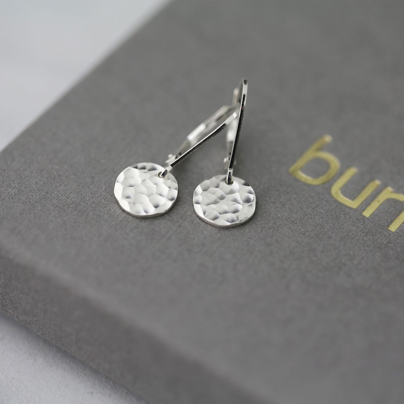 May include: A pair of silver hammered disc earrings. The earrings have a textured surface and are suspended from lever-back closures. The earrings are displayed on a gray surface.