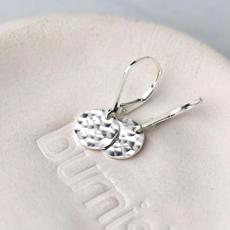 May include: Silver hammered disc earrings with lever-back closures. The earrings have a textured, dimpled surface and are displayed on a white surface. The earrings are a simple, elegant design.