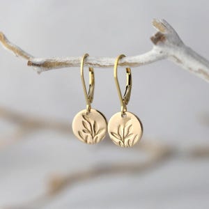 May include: A pair of gold-colored leverback earrings. Each earring features a small, round charm with an embossed leaf design. The earrings are hanging from a white branch against a soft, blurred background.