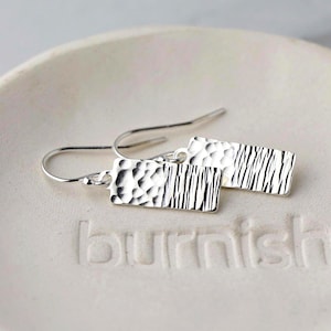May include: A pair of silver rectangular earrings with textured surfaces. One earring has a hammered texture, the other has vertical lines. The earrings hang from curved ear wires. The earrings are on a white surface.