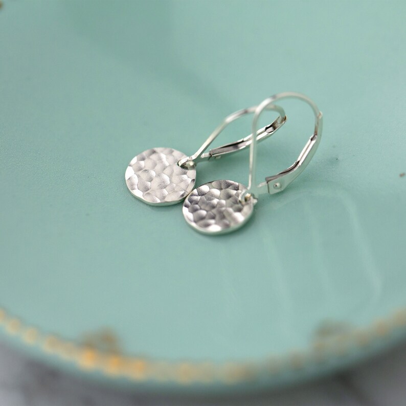 May include: Silver hammered disc earrings with lever-back closures. The earrings feature a textured, hammered surface on the circular pendants. The earrings are displayed on a light teal surface.