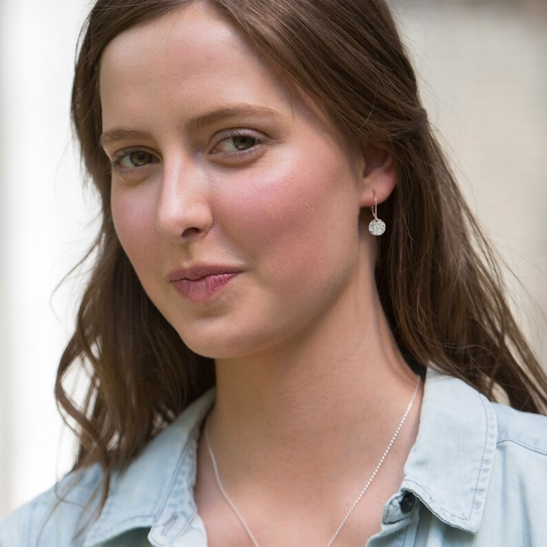 May include: A woman wearing a light blue denim shirt and a silver necklace. She is also wearing a pair of small, round, sparkly earrings. The earrings are a focal point, catching the light and adding a touch of elegance to the look.