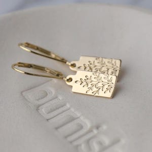 May include: A pair of gold-colored earrings. Each earring features a rectangular tag with an etched botanical design. The earrings have a lever-back closure. The earrings are displayed on a white surface.