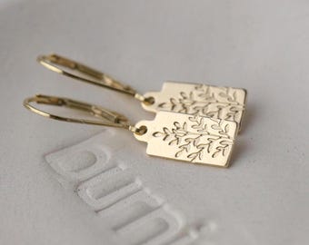 May include: A pair of gold-colored earrings. Each earring features a rectangular tag with an etched botanical design. The earrings have a lever-back closure. The earrings are displayed on a white surface.