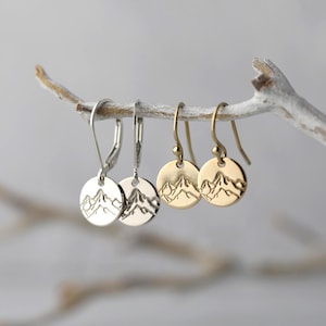 May include: Four pairs of earrings with a mountain design are displayed on a branch. Two pairs are silver-toned, and two are gold-toned. Each earring features a circular pendant with an engraved mountain range.