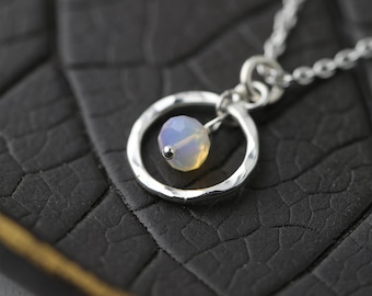 May include: A delicate silver necklace featuring a small, faceted, opalescent gemstone pendant. The pendant is suspended within a hammered silver circle, attached to a fine chain. The gemstone has a soft, iridescent glow.