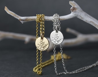 May include: Two delicate necklaces with round pendants, one gold-toned and one silver-toned, each featuring an engraved floral design. The necklaces hang from a white branch against a dark gray background, showcasing their intricate details and elegant simplicity.