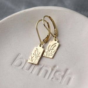 May include: A pair of gold-colored leverback earrings. Each earring features a rectangular tag with an etched botanical design of leaves and stems. The earrings are displayed on a white, textured surface.