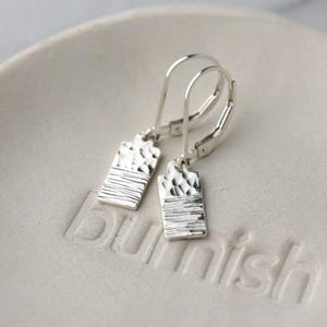 May include: A pair of silver rectangular earrings with textured surfaces. The top half of each earring has a hammered texture, while the bottom half features horizontal lines. The earrings are displayed on a white ceramic surface.